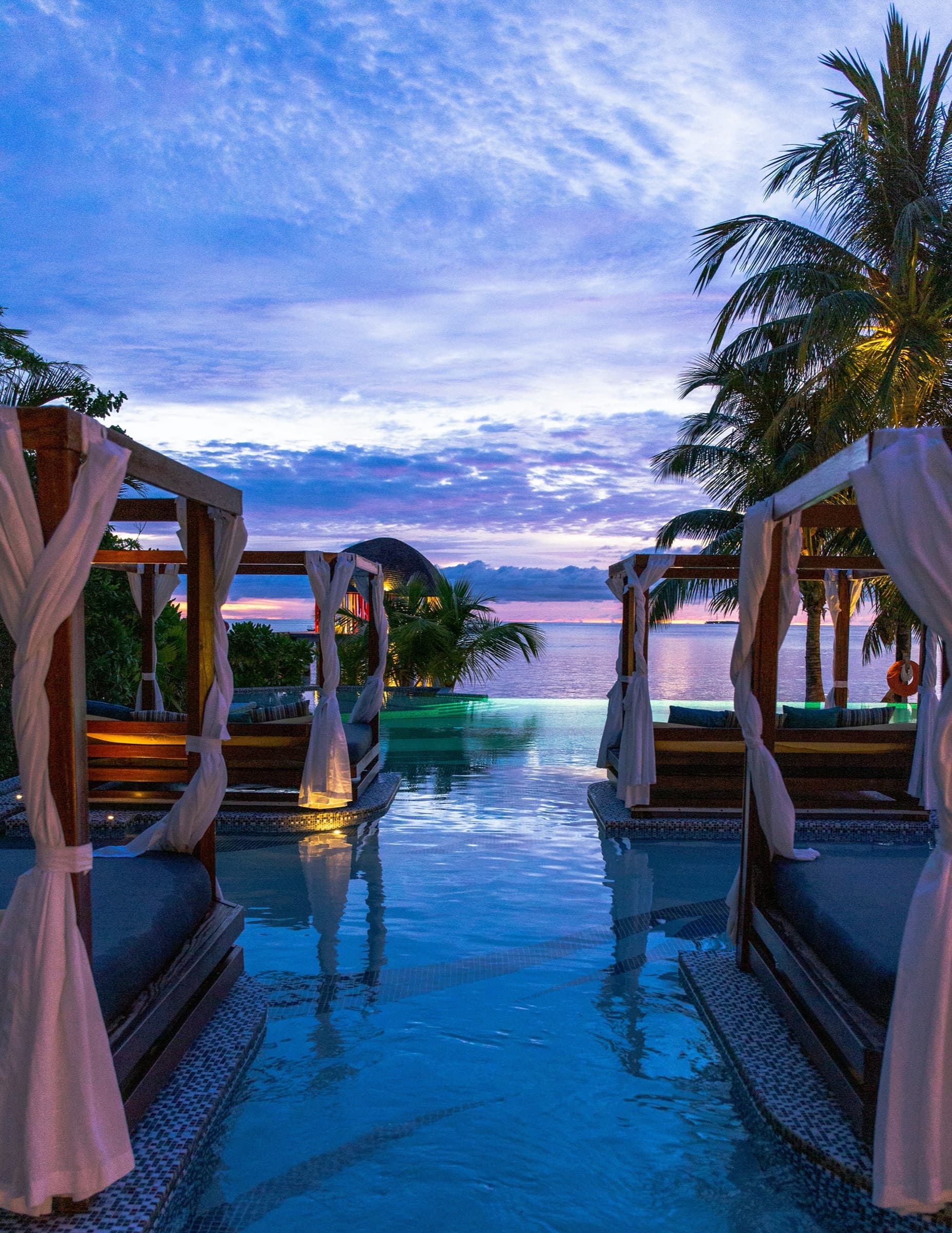 Canopied poolside daybeds with white drapes beside a resort infinity pool under a violet dusk sky