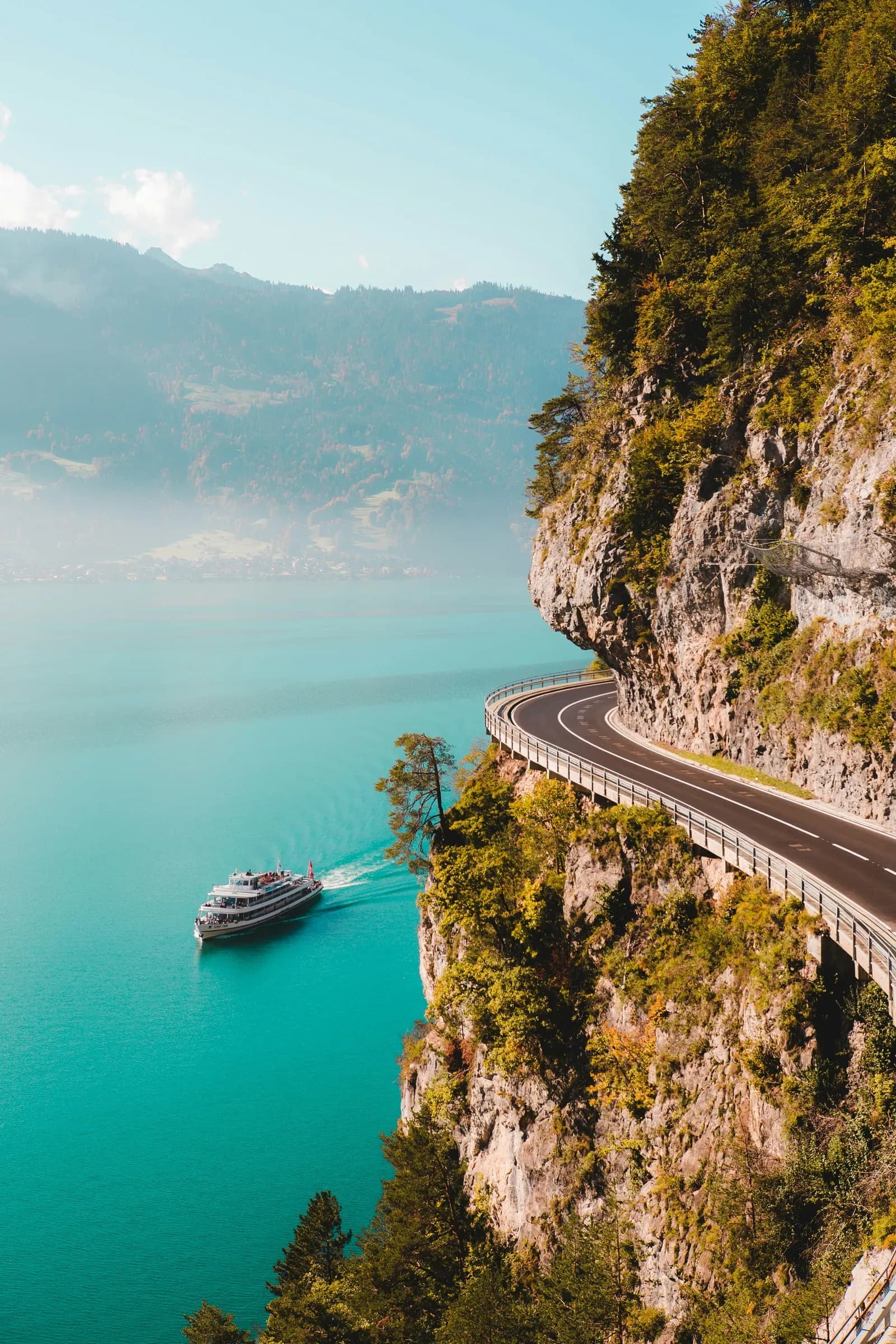 A road cut into a sea cliff above turquoise water, with a boat passing below