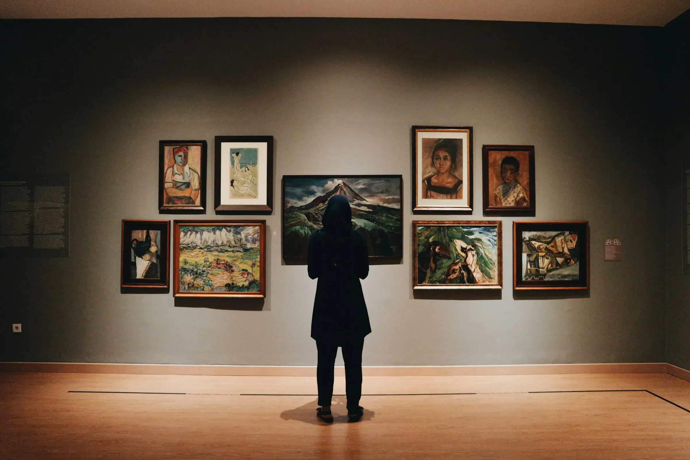 A visitor standing before a wall of framed paintings in a dim gallery