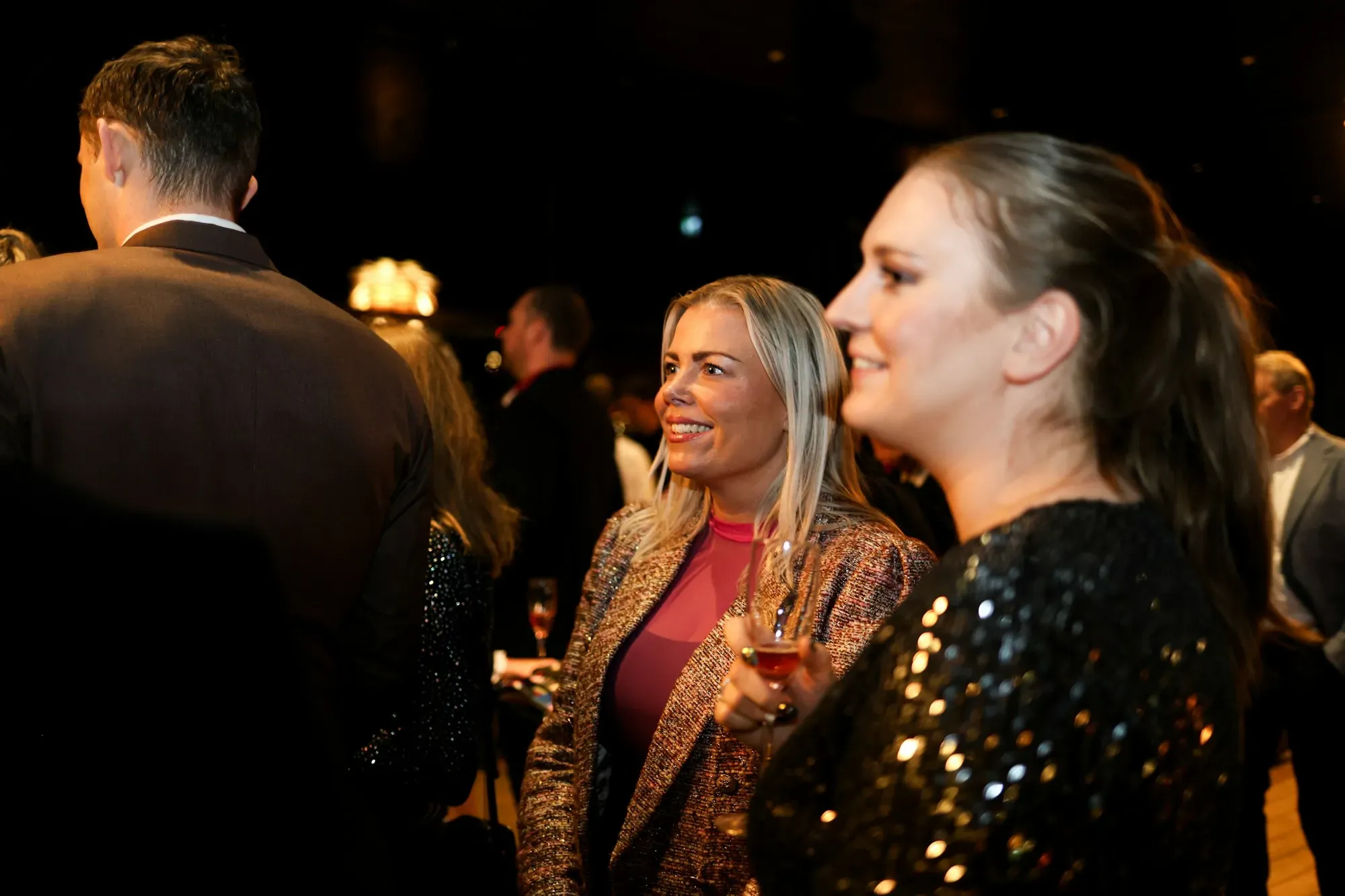 Two women in evening dress laughing among guests at a night event