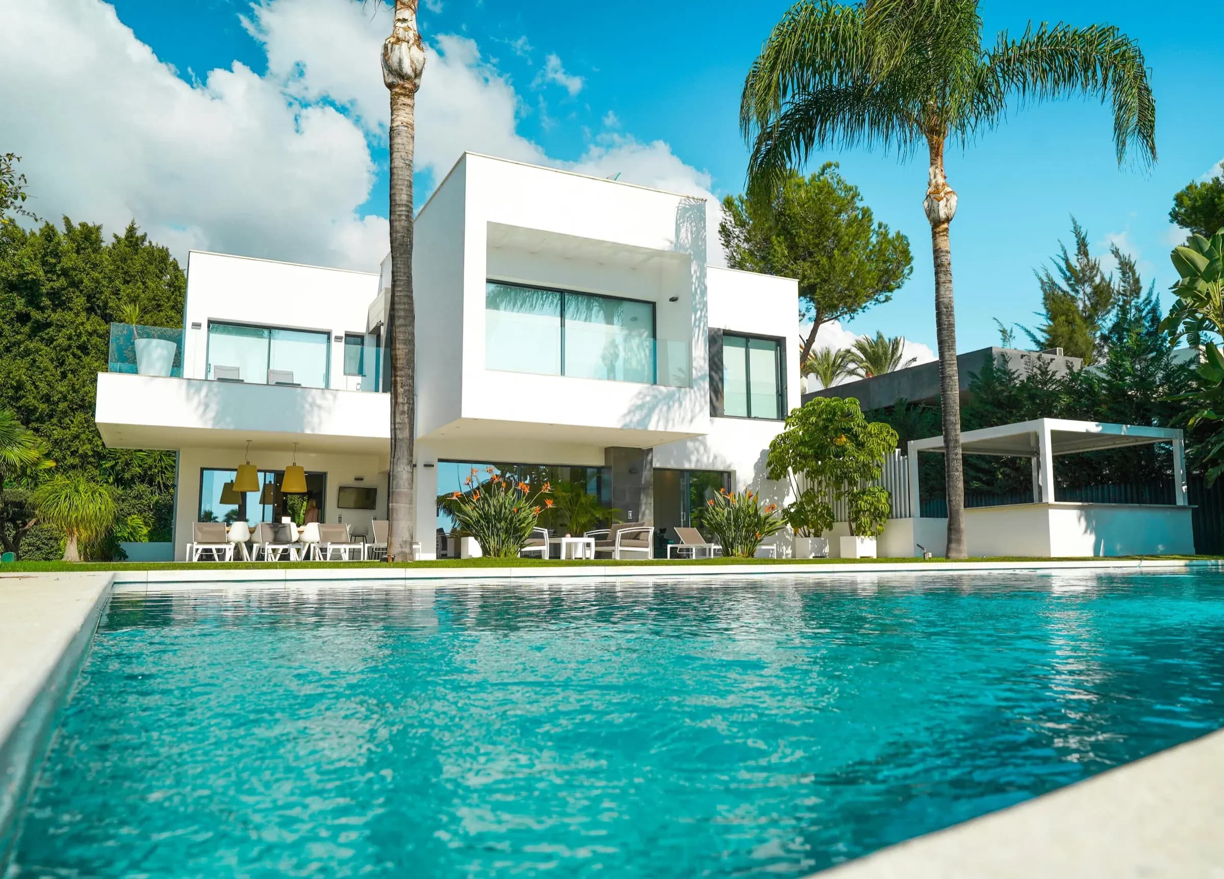 A white modern villa behind a turquoise pool and palm trees